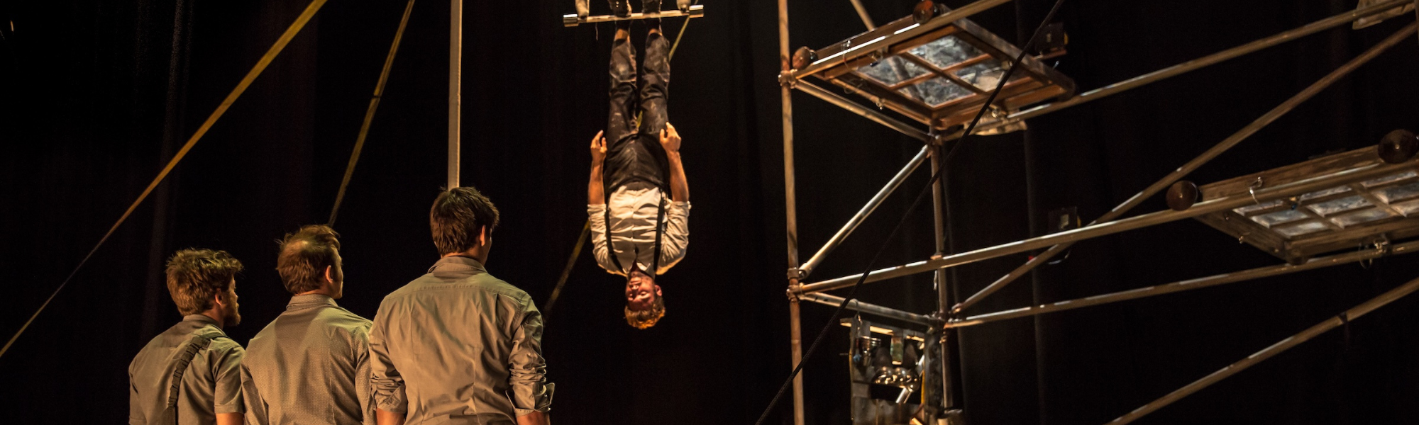 person suspended upside down on a scaffolding frame with 3 bystanders looking on